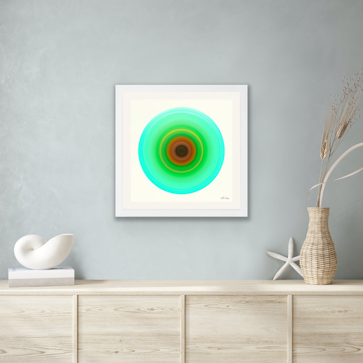 A framed square wall art print hangs on a light gray wall above a pale wood console. The artwork shows concentric circular bands in green, turquoise, and brown on a white background.